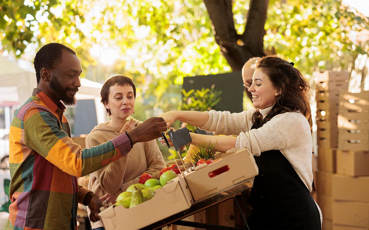 Multiracial couple tasting organic produce at a farmers market while talking to a woman selling the produce..